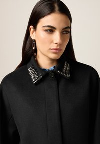 Abrigo negro de lana con un cuello estructurado que presenta adornos metálicos. La textura es suave con detalles visibles de costura.