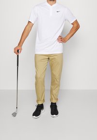 Bílá polo košile s krátkým rukávem a knoflíkovým límcem, béžové chino kalhoty a černé tenisky s bílými podrážkami, držící golfové hole.