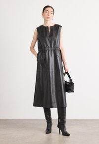 Schwarzes ledernes Maxikleid mit ärmellosem Design, elastischem Bund, tiefem Ausschnitt und seitlichen Taschen. Kombiniert mit knielangen schwarzen spitzen Stiefeln.