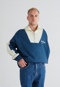 Pullover blu navy con colletto bianco e chiusura a mezza zip, con una vestibilità ampia e testo ricamato sul petto. Indossato con jeans in denim.