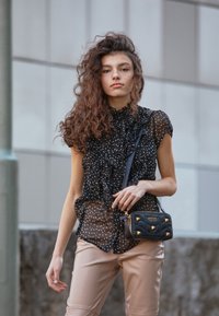 Černá blůza s puntíky a volány, průsvitná textura; kombinováno s béžovými úzkými kalhotami a malou černou prošívanou kabelkou se zlatými akcenty.