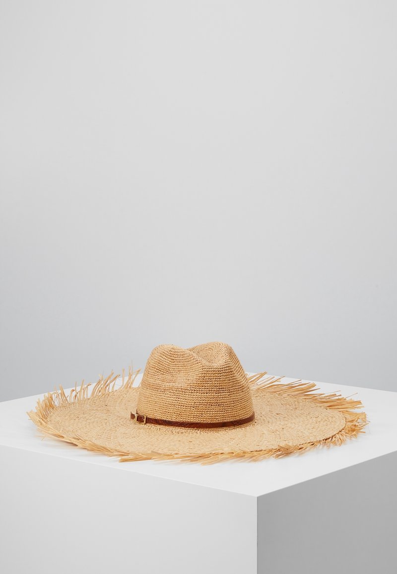 Chapeau en paille naturelle à large bord avec des bords effilochés et un bandeau en cuir marron, exposé sur un piédestal blanc devant un fond gris uni.