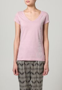 Camiseta de algodón rosa con escote en V y mangas cortas, con texto brillante en el lateral. Combinada con pantalones estampados en blanco y negro.