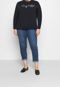Zwart sweatshirt met witte "Tommy Hilfiger" logo, gecombineerd met opgerolde spijkerbroek in het blauw en zwarte instappers. Eenvoudige, casual outfit met strakke lijnen.