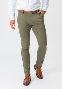 Oliven grønne strækbukser med slim fit, flad front, sidelommer og brunt læderbælte, parret med skinnende brune dressko.