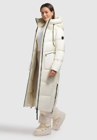 Cappotto lungo in piumino di colore crema con un cappuccio voluminoso, dotato di cerniere nere, tasche laterali e un design trapuntato per l'isolamento.