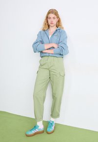 Svetlomodrá mikina na zips s logom, zelené cargo nohavice a multikolorové tenisky s tyrkysovými, ružovými a bielymi akcentmi. Model stojí na zelenej podlahe.