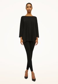 Sort tunika med løs pasform, rund halsudskæring og trekvartlange ærmer, kombineret med sorte leggings og spidse hæle. Glat tekstur.