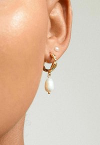 Gul hoops ørestikker med et tekstureret design, der har en hængende hvid perle. Lille perle stud ørestik vises i den øverste del af øret.