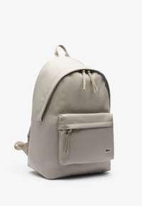 Beżowy plecak z przednią kieszenią na zamek błyskawiczny, podwójnymi suwakami, górnym uchwytem, szelkami oraz małym zielonym logo krokodyla na kieszeni.