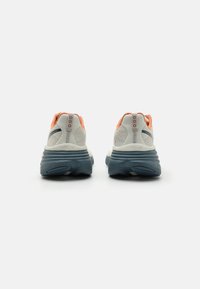 Baskets de course blanches et orange avec un dessus texturé, semelle rembourrée bleu marine et un design de talon prononcé. Présente une étiquette de marque sur le talon.