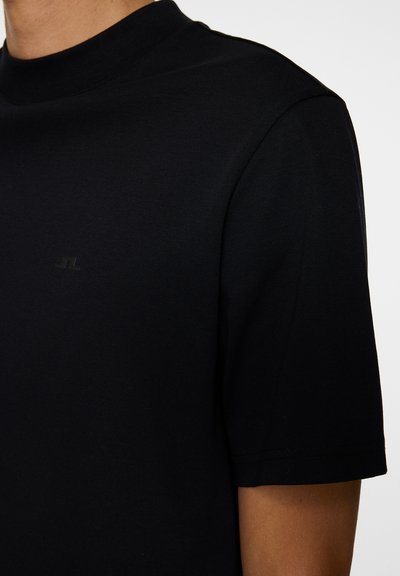 Persona che indossa una semplice maglietta nera a maniche corte con un sottile logo piccolo sul petto, mostrata dalla scollatura a metà torso.