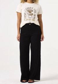 Camiseta gráfica de crema con diseño floral, combinada con pantalones de pierna ancha texturizados en negro. Sandalias negras simples visibles en los pies.