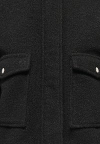 Manteau en laine noire avec un tissu texturé, deux poches avant avec des boutons blancs, et une silhouette droite. Ouverture unique à l'avant.