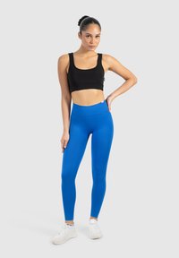 Soutien-gorge de sport noir avec des bretelles larges et un design court, associé à un legging bleu taille haute et ajusté, et des chaussures de sport blanches.