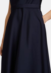 Robe bleu marine présentant une texture satinée lisse, un corsage ajusté, une jupe évasée et une ceinture coordonnée ; comprend un détail de poche latérale.