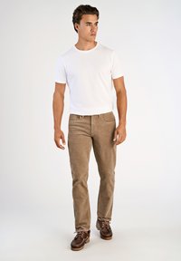 Biała dopasowana koszulka, jasnobrązowe spodnie z sztruksu i brązowe sznurowane buty. Model stoi, ręce w luźnej pozycji, neutralne tło.