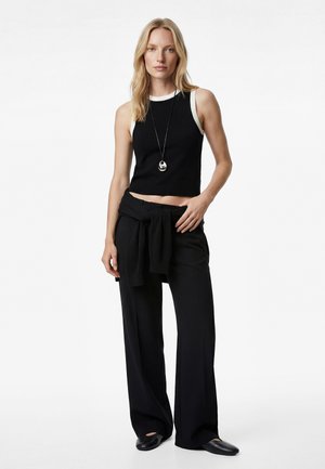 Mujer rubia con top corto sin mangas en blanco y negro, pantalones negros de pierna ancha con suéter atado a la cintura, zapatos planos negros y collar con colgante.