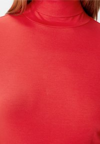 Close-up van een persoon die een rode lange mouw turtleneck shirt draagt, met zichtbare textuur van de stof en details van de halslijn.