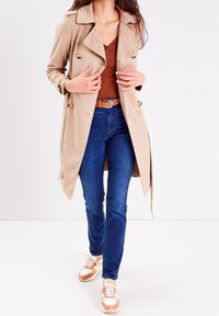 Beige dubbelzijdige trenchcoat met een ingesneden kraag, knopen, gedragen over een bruine top en blue jeans, gecombineerd met crème en oranje sneakers.