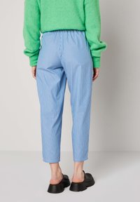 Inimene, kes kannab helesiniseid ja valge triibulisi lühikesi pükse, rohelist pikkade varrukatega kampsunit ning musti libisev jalanõusid, seisab ilma taustata tausta ees.