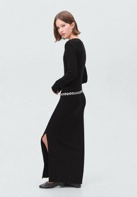 Robe maxi noire à côtes avec manches longues, présentant une fente sur le côté et une ceinture accent circulaire en argent. Texture lisse, silhouette près du corps.