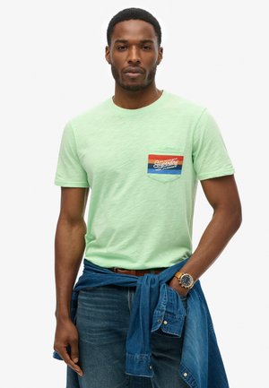 Camiseta de manga corta verde menta con un pequeño bolsillo que presenta un diseño a rayas en azul y naranja. Llevada con jeans de mezclilla y una camisa de mezclilla atada.