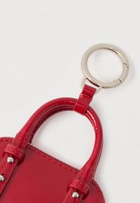 Uchwyt czerwonej lakierowanej torebki przymocowany do srebrnego metalowego breloczka na białym tle.