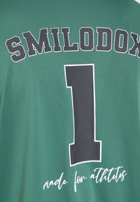 Zelený sportovní dres s nápisem "SMILODOX" v tučných písmenech, velkým číslem 1 a nápisem "vyrobeno pro sportovce" napsaným kurzívou.