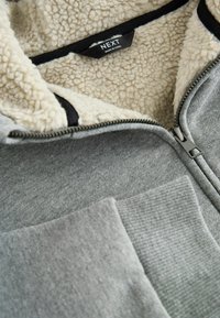 Grå sweatshirt med en fleece-fodrad krage, med dragkedja och en texturerad ficka. Svart etikett synlig invändigt.