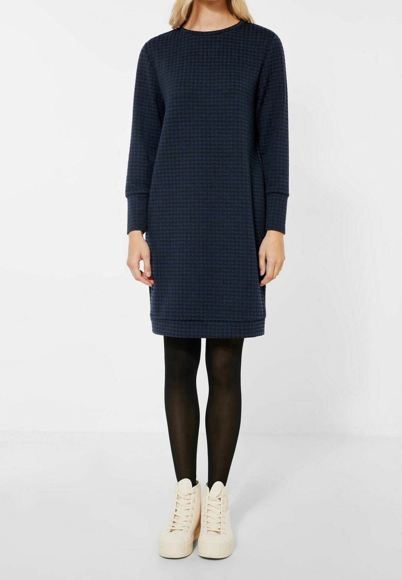Robe tricotée bleu marine à manches longues, col rond et motif texturé. Associée à des collants noirs et des baskets montantes crème.