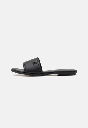 Sort flad slid sandal med bred rem med lille præget logo på en ensfarvet hvid baggrund.
