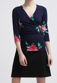 Marinblå omslagstop med trekvartslång blommig ärm som har rosa och gröna blommor, kombinerad med en strukturerad svart kjol.