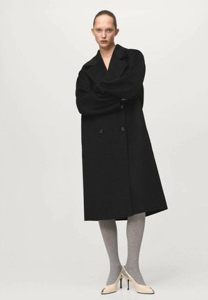 Czarny płaszcz dwurzędowy z szerokimi klapami, o teksturze wełny, długości do kolan, zestawiony z szarymi rajstopami i beżowymi butami na wysokim obcasie.