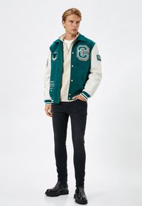 Teal ja cream värvi varsity jope, millel on tikitud plaastrid, koos beez jailbreakiga ja mustade kitsaste teksadega, lõpetatud mustade nahkadega.