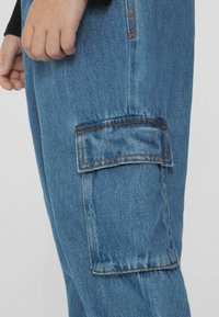 Modré denimové cargo kalhoty s kapsou na boku, oranžovým prošíváním a volným střihem. Látka má mírně texturovaný povrch.