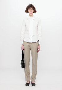 Camicia bianca con buttons, cravatta, pantaloni beige, scarpe flat nere e borsa nera, in piedi davanti a uno sfondo bianco.