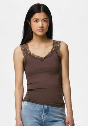 Young woman with long dark hair wearing a fitted brown tank top with lace trim and light blue jeans, standing against a plain background.