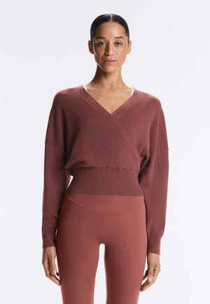 Frau mit dunklem Haar zu einem tiefen Dutt gebunden, trägt einen langärmeligen, gerippten Wickelpullover und hoch taillierte Leggings, beide in gedämpften rötlich-braunen Tönen.