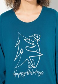 Türkises Langarmshirt mit einem weißen Grafikelement, das einen Bären mit einem Weihnachtsmannhut zeigt, der einen Weihnachtsbaum hält, mit dem Text "Frohe Feiertage".