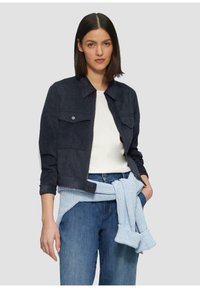 Vrouw in een marineblauwe jas over een witte top, met een lichtblauwe trui om haar middel en blauwe jeans, staande tegen een effen achtergrond.