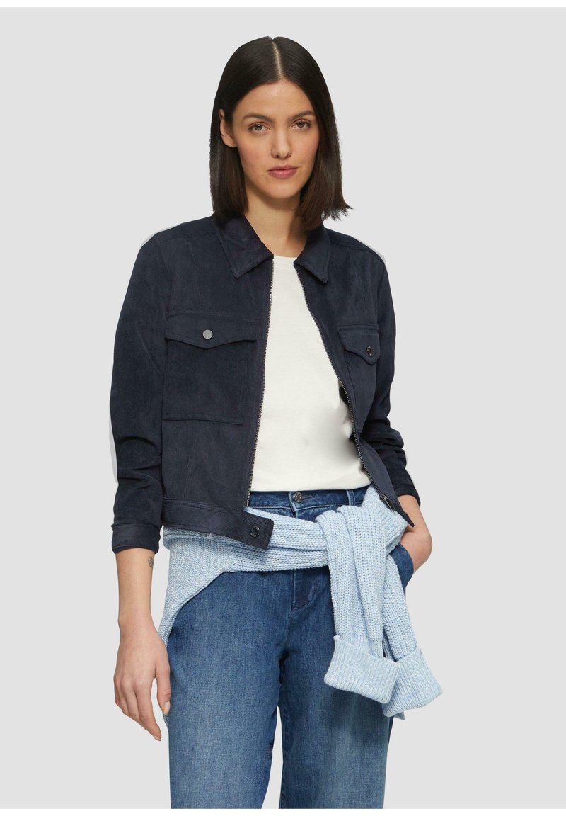 Vrouw in een marineblauwe jas over een witte top, met een lichtblauwe trui om haar middel en blauwe jeans, staande tegen een effen achtergrond.