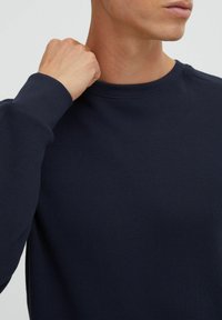 Tricou bleumarin dintr-un material texturat, cu model de plasă. Dispune de un guler rotund și manșete cu nervuri, fiind parțial arătat cu o mână care apucă gulerul.