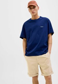 Tamsiai mėlynas medvilnės T-shirt su mažu baltu "Originals" logotipu, derinamas su smėlinėmis šortais, turinčiais sukti kraštą, kad būtų patogiai prigludę.