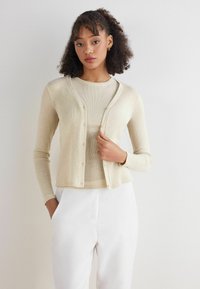 Jasno-beżowy kardigan z żebrowanej tkaniny, z okrągłym dekoltem, długimi rękawami i zapięciem na sześć guzików, nałożony na dopasowaną bluzkę.