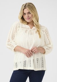 Light cream blouse with intricate eyelet pattern, slightly gathered bodice, round neckline, and 3/4 sleeves, paired with dark jeans.