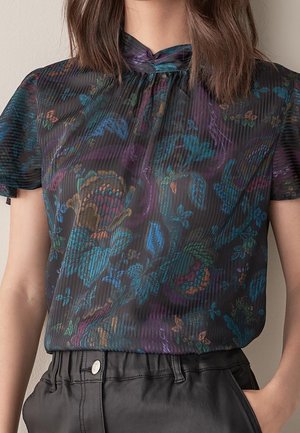 Vrouw draagt een doorschijnende zwarte top met korte mouwen, kleurrijke bloemenpatronen en een hoge hals, gecombineerd met zwarte leren broek.