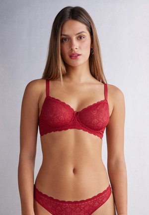 Red lace bra with scalloped edges and adjustable straps, featuring a floral pattern and a central bow detail. Matching underwear visible.