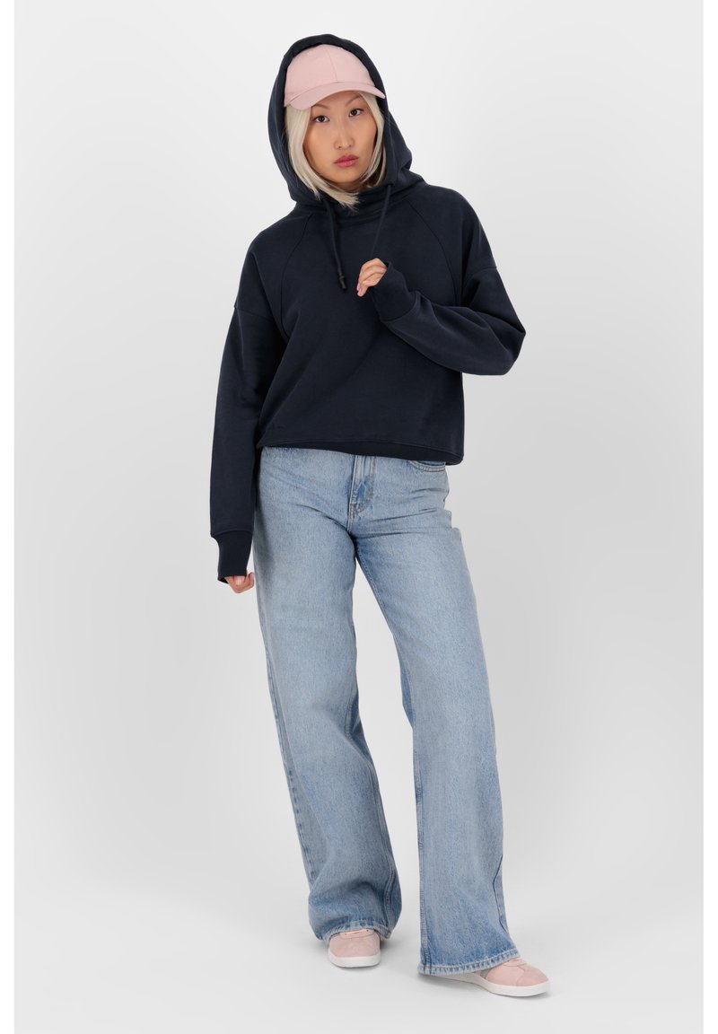 Felpa cropped blu marino con cappuccio regolabile, abbinata a jeans a gamba larga blu chiaro e sneakers rosa pallido, su uno sfondo bianco.