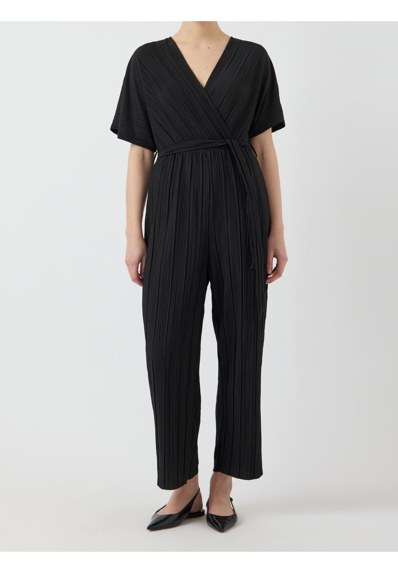 Femme portant une combinaison cache-cœur plissée noire à manches courtes et ceinture à la taille, associée à des chaussures slingback noires à bout pointu.